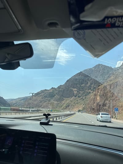 The image is taken from inside a vehicle, looking forward through the windshield. The road ahead curves to the left, with a white car in the right lane. The right side of the road is bordered by a steep, rocky mountain face. The left side of the road has a barrier, beyond which are train tracks with several freight cars. The sky is blue with some clouds. A dashcam is visible on the dashboard, and a box of tissues is hanging from the rearview mirror. A road sign indicating a U-turn in 250 meters is visible on the right side of the road. The interior of the car shows part of the dashboard and infotainment screen. The scene appears to be a highway or main road passing through a mountainous or hilly region, potentially in or near the city of Beshkul, Uzbekistan.