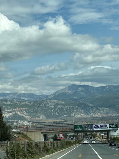 The image presents an outdoor scene depicting a multi-lane highway in the city of Menteşe, Turkey, under a partially cloudy sky. The highway, marked with white lines and a yellow directional arrow, extends towards the viewer and then under a concrete overpass. Multiple vehicles, primarily white cars and at least one white bus, are observed traveling away from the viewer on the highway.
In the background, a range of rocky mountains, partially covered with sparse green vegetation, dominates the horizon. Several wind turbines, approximately six, are visible atop or along the ridges of these mountains. On the lower slopes and in the valley between the mountains and the foreground, an urban development consistent with Menteşe is visible.
In the midground, a concrete overpass spans horizontally across the highway. Attached to this overpass are large green billboards displaying Turkish advertisements, including "EKŞİOĞLU," "%0 FAİZ," and "990.000 TL." To the right of the highway, before the overpass, a red and white triangular road sign indicating falling rocks is visible. The immediate left foreground features a dark green coniferous tree and a metal fence lined with green shrubs. No individuals are explicitly depicted in the scene.