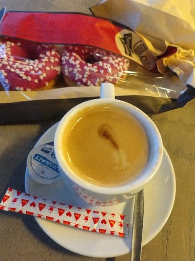 An overhead shot shows a coffee cup on a saucer with a spoon and sugar packet. Next to it is a bag of pink donuts with white heart-shaped sprinkles. The coffee has a light brown hue with a small darker brown spot in the center. The sugar packet has a repeating pattern of red triangles and hearts. A small container is under the coffee cup. The donuts are in a clear plastic bag within a brown paper bag.