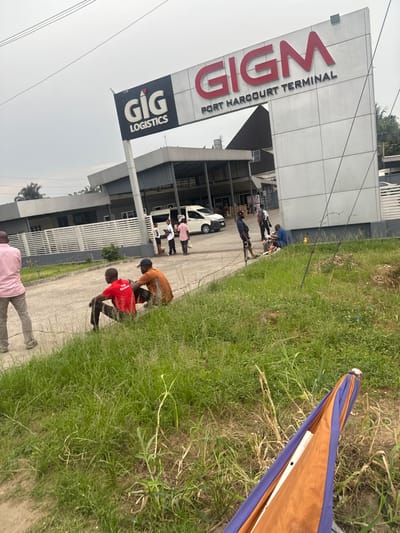 The image depicts GIG Logistics and Port Harcourt Terminal building. Several people are gathered outside the building's entrance. A white car is parked nearby. The foreground shows a grassy area with an individual and a broken orange and blue umbrella. The image is taken at an angle, causing the building and surroundings to appear tilted.
