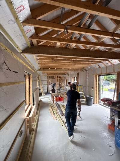 The image displays the interior of a building under construction in Skipsea, United Kingdom. The structure features exposed timber roof trusses, ceiling joists, and wall framing. Walls are partially insulated with silver-faced 'Recticel' thermal panels and framed for interior finishing. The floor is an unfinished concrete slab.
Four individuals are present. One person in the foreground, seen from behind, walks towards the center of the room wearing a dark shirt and jeans. Three other workers are observed in the mid-ground: one is bent over, one stands near a table with two orange buckets, and another is positioned by a workbench.
Various construction materials are visible, including stacks of timber battens and planks on the floor, and large white panels (likely drywall) leaning against the left wall. Equipment includes a yellow step ladder, a black trash bin, a blue barrel, and multiple power tools situated on work surfaces. Exposed electrical wiring runs throughout the timber framework. An open doorway on the right reveals an exterior paved area, green trees, and a dark-colored vehicle, indicating the site's surroundings.