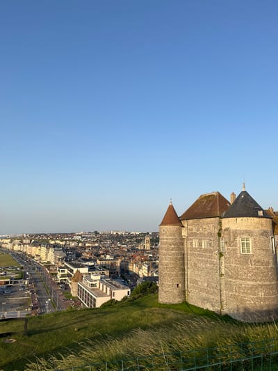 The image shows a panoramic view of Dieppe, France, from a high vantage point. Dominating the foreground is a section of a stone castle, with a conical roof and several windows visible. To the right, a steep grassy slope, dotted with tall, dry grass and a green metal fence, descends towards a dense urban landscape. The town stretches out horizontally across the middle ground, featuring a variety of buildings with pitched roofs and a coastline with a promenade lined with cars.
The scene appears to be set during a clear day, likely late afternoon or early evening, given the warm, soft light casting shadows and illuminating the town. The sky is a vibrant, cloudless blue, transitioning to a lighter hue near the horizon. The overall atmosphere is serene and picturesque, showcasing the historical architecture of the castle against the backdrop of a bustling coastal town. There is no visible text in the image.