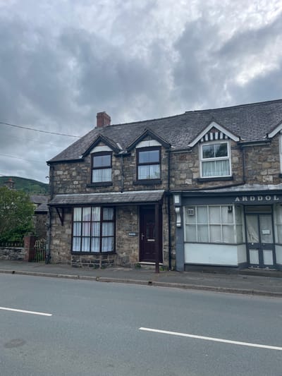The image shows a view of a street in Llangynog, United Kingdom, with a row of stone-built terraced houses and a shop. The sky is overcast.
The property on the left is a two-story house with a dark tiled roof and a brick chimney. The ground floor features a large bay window with white curtains, and a dark wooden door. Above the bay window, there is a smaller window, also with white curtains. The building appears to be constructed of rough-hewn stone blocks. A small sign above the door reads "Gwynfa Dale." A low stone wall and a metal gate are visible to the left of the house, with some greenery behind them.
To the right of the house is a commercial property, likely a shop, with a dark grey facade. The shop has a large display window with white panelling and visible white curtains behind the glass. Above the window, the word "ARDDOL" is visible in white lettering against the dark grey background. There are double doors on the right side of the shop front.
The street in front of the buildings is paved, with a white line painted down the center. A concrete curb separates the road from the pavement. In the background, on the left side, a green hillside is visible under the cloudy sky. Power lines run across the top of the image.