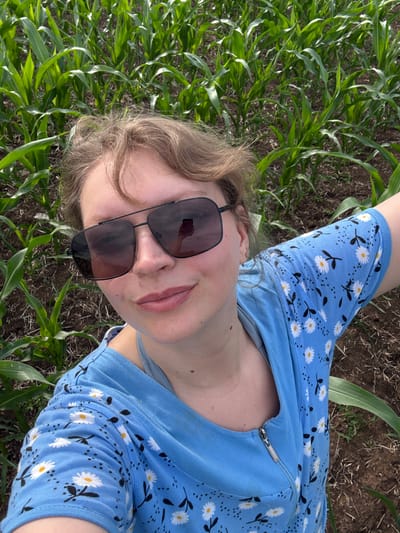 The media file shows a person lying down in a cornfield in Merinovshchina, Russia. The person is wearing a blue shirt with a daisy pattern and black sunglasses. The corn stalks are green and vibrant, suggesting it is daytime and likely summer. The ground appears to be dry soil, possibly indicating a warm and sunny day. The perspective is a close-up, angled shot, with the person's face and upper body filling most of the frame. The person's expression is neutral, and their gaze is directed upwards, away from the camera. The scene is natural and appears to be a candid shot. There is no visible text in the image.