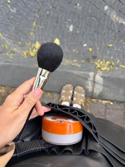 A first-person, downward-facing shot captures a left hand holding a black makeup brush with a silver ferrule. A small ring is visible on the hand's ring finger. Below the hand, a black faux leather makeup organizer holds a clear container with an orange lid, identified as Coty Airspun loose face powder. In the background, blurred, a person's feet are visible, wearing tan sandals over white socks, standing on a grey cobblestone surface littered with scattered yellow and brown leaves. The specific location within West Jerusalem, Israel, is not discernible from the visual evidence.