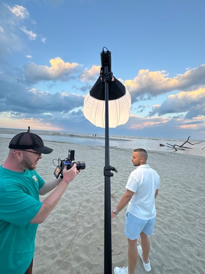 Two men are on a beach. One man in a green t-shirt and black baseball cap is holding a camera and filming the other man. The man being filmed is wearing a white polo shirt, blue shorts, and white sneakers. A large white light diffuser is positioned on a stand between the photographer and the subject. The beach is sandy, and there are some branches lying on the sand in the background. The sky is cloudy with patches of blue. The location is likely Novo Oriahovo, Bulgaria, based on the presence of a beach.