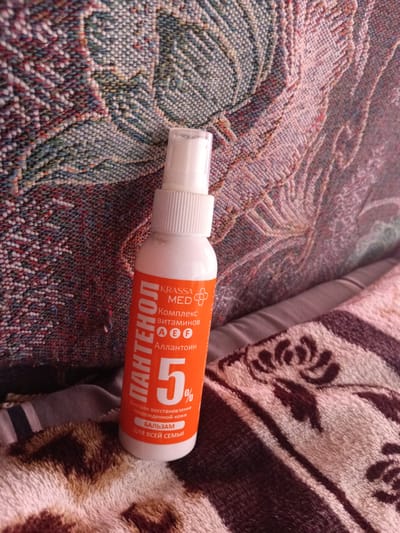 A partially visible white spray bottle with an orange label sits on a woven blanket with a muted color palette. The label prominently features the Cyrillic text "ПАНТЕНОЛ" (PANTENOL) vertically, along with "KRASSA MED" and "Комплекс витаминов" (Vitamin Complex). Below this, "АЕF" and "АЛЛАНТОИН" (Allantoin) are visible. A large "5%" graphic is also present, followed by smaller text indicating "Быстрое восстановление поврежденной кожи" (Rapid restoration of damaged skin) and "БАЛЬЗАМ ДЛЯ ВСЕЙ СЕМЬИ" (Balm for the whole family). The bottle is positioned slightly to the right of the center of the frame, angled slightly forward. The background consists of the textured fabric of the blanket. There are no people or discernible city elements present in the image.