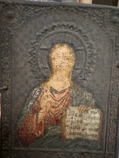 The image shows an aged religious icon, possibly Orthodox, depicting a figure with a halo holding a book. The icon has a dark, weathered appearance with visible wear and deterioration of the paint. The figure is wearing robes and holding a book with script, though the script is heavily degraded. The icon is encased in what appears to be a dark, decorative border. The location in Sayansk, Russia, is based on external information, and does not appear in the image itself.