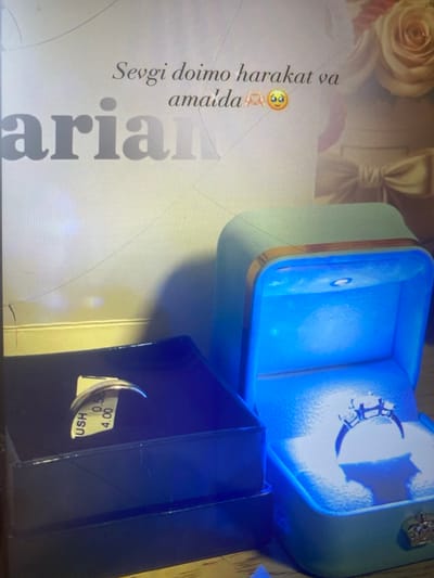 The image depicts two jewelry boxes, one open and illuminated with blue light, showcasing a diamond ring. The second box is closed and dark, with a tag visible on the side of a ring inside. The background features text that appears to be "Sevgi doimo harakat va amaida" followed by emojis, and a large blurred word "arian". A bouquet of flowers is also visible in the background. There is no discernible information about the city of Arbagish, Uzbekistan, in the image.