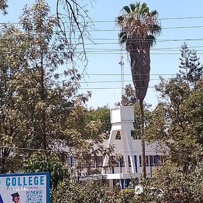 The image depicts an outdoor, daytime scene within Machakos Central ward, Kenya, characterized by a mix of dense vegetation, architectural structures, and utility infrastructure under a clear light blue sky.
In the foreground, a partial blue sign is visible in the lower-left corner, displaying "O COLLEGE" and "Beyond Training..." along with a QR code and a small monochrome image of a person in a graduation cap.
The midground is dominated by lush green foliage from numerous trees and bushes. A prominent, tall palm tree with a thick, textured trunk and a crown of green fronds stands towards the right of center. Behind the vegetation, a multi-story white building with blue vertical trim, possibly outlining windows or structural elements, is partially visible. Directly in front of this building, a distinct white, multi-level architectural structure features an inverted trapezoidal base with triangular voids, supporting a rectangular upper section.
In the background, a tall, slender metal communication tower equipped with multiple antennae rises behind the palm tree and white structure. Multiple dark, horizontal utility lines traverse the upper and mid-portions of the frame. No individuals are visible.