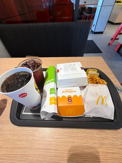 On a wooden table, a black plastic tray holds a fast-food meal. In the foreground, a white paper cup with a Coca-Cola logo is filled with a dark beverage and ice. Behind it, a clear glass contains a dark red beverage with ice and visible dark fruit pieces. To the right of the beverages, a white and green cardboard container is partially visible. Next to this, a white box with "40" printed on it sits on top of another white box. To the right of these boxes, a small yellow sauce container is positioned above a portion of waffle fries. Below the fries is a white paper bag with a prominent yellow McDonald's logo. In front of the fries and sauce, an orange box with "Chicken McNuggets" and the number "6" printed on it is visible. The background shows a dark upholstered booth and glimpses of a fast-food restaurant interior with red and grey accents. The image appears to be taken in a McDonald's restaurant.