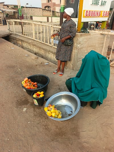 Two women are on a street. One stands next to a cooking gas sign, wearing a headscarf and patterned dress. She holds something in her hand. The other woman squats, covered entirely by a green cloth. Three containers of cashew apples are displayed: a large black tub, a small black bucket, and a metal bowl. The ground is paved. A white metal railing lines a raised sidewalk.