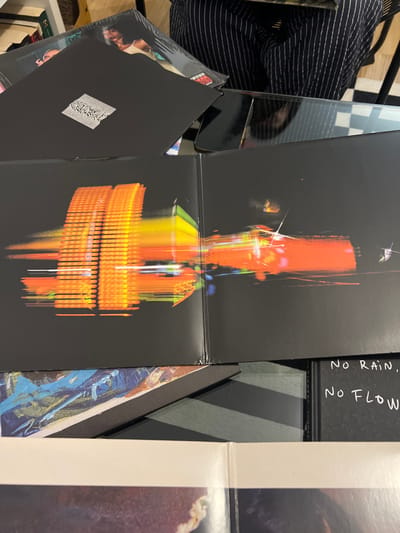 The image shows a collection of vinyl records. Open in the center, one album features a black cover with a distorted, colorful image resembling an abstract structure. To the left, another album bears a black cover with a smaller, geometric white design. "NO RAIN. NO FLOW." is printed on a black album spine. Various other album covers are partially visible, including one with a striped pattern and another with a predominantly white and brown color scheme. A striped garment lies in the background.