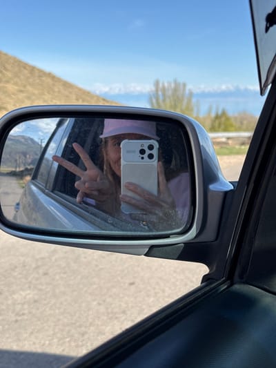 The image shows a selfie taken in the side mirror of a car. The reflection reveals a person holding a smartphone, making a peace sign. Outside the car window, the landscape includes a body of water (likely Lake Issyk-Kul), green hills, and a clear blue sky. Based on the landscape and water body, the location is likely near Cholpon Ata, Kyrgyzstan. The car is traveling on a road.