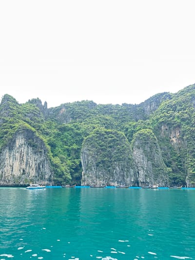 The image depicts a view of a body of turquoise water with a line of traditional longtail boats docked along a floating blue barrier. In the background, steep, verdant limestone cliffs rise from the water's edge. A white speedboat with a blue stripe is seen in the middle of the water, moving from left to right. The sky above is overcast and white. This scene is typical of the coastal scenery found in areas like Haad Yao, Thailand, known for its limestone karst formations and clear waters.