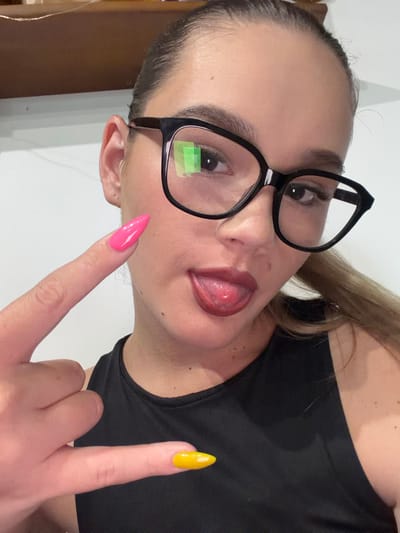 In this media file, a close-up shot features a young woman, likely in her late teens or early twenties, with fair skin and light brown hair pulled back. She is wearing black-rimmed glasses and a black top. Her tongue is playfully sticking out, and her eyes are looking towards the camera. Her left hand is raised, with a bright pink, pointed fingernail prominently displayed, and her index finger is directed towards the camera. Her right hand is also visible, with a yellow fingernail.

The background appears to be an indoor setting with a white wall. There are no discernible cues about the time of day or weather conditions. The overall mood conveyed is playful and perhaps a bit flirtatious. There is no visible text in the image. Given the provided location context of Beguntsi, Bulgaria, it is possible this is where the photo was taken.