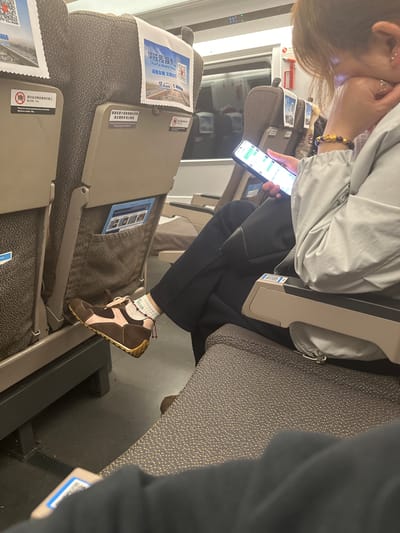 An indoor, eye-level shot captures a person seated on what appears to be a train or high-speed rail in Yuhang District, China, engrossed in their smartphone. The person has light brown hair and is wearing a light grey jacket, black pants, white socks, and distinctive brown and pink shoes. A gold bracelet adorns their right wrist, and a red beaded anklet is visible on their left ankle above the white sock, which features the text "min".

The individual is looking down at their phone, held in their hands, with their left hand positioned near their face. The phone's screen displays what looks like a messaging application or perhaps financial data with various numbers.

The setting is a public transport carriage with multiple rows of seats upholstered in a dark, textured fabric with small gold or light-colored flecks. Foldable tray tables are attached to the back of the seats. On the back of the seat in front, a white cover displays Chinese text for "Hua Tie Media Group" and "诚信友善文明出行" (Integrity, Friendliness, Civility, Travel) alongside QR codes. Another sticker warns, "请勿坐或搁放重物在桌上 最大负重 10kg" (Please do not sit or place heavy objects on the table, maximum weight 10kg). Another sign indicates, "插座设置于座垫接缝处前端 请正确使用防止触电" (Socket is located at the front of the seat cushion joint, please use correctly to prevent electric shock). A digital display on the wall shows "11", possibly indicating temperature or time. The lighting is artificial, suggesting an indoor environment, and the person appears relaxed and focused on their device.