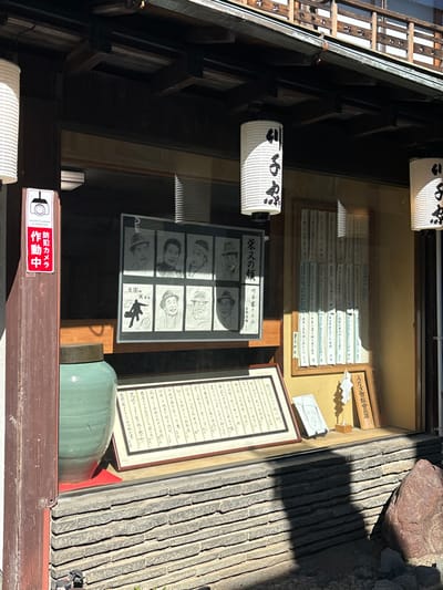 The image shows a storefront window with a sign in Japanese, "Photography Prohibited," and a display of framed artwork. The artwork features black and white portraits of seven men. Two white paper lanterns are visible above the display. There is a ceramic pot in the window. The image appears to have been captured in front of a building at the coordinate location (latitude: 35.758, longitude: 139.877) in Katsushika City, Japan.