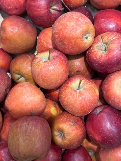 The image is a close-up, overhead view of a pile of ripe apples. The apples vary in color from deep red to a lighter reddish-orange, with some showing yellow undertones and small, light-colored speckles on their skin. Most of the apples are round, though some are slightly irregular in shape. Stems are visible on a few of the apples, protruding from indentations at the top. The apples are tightly packed together, suggesting they are either for sale or have been freshly harvested. The lighting appears to be artificial or overcast, casting a soft, diffused light on the fruit. There is no visible text in the image. The setting is likely a market stall or grocery store display, given the abundance and presentation of the fruit. The scene is focused entirely on the apples, with no other objects or living beings visible. The image conveys a sense of freshness and abundance of produce.