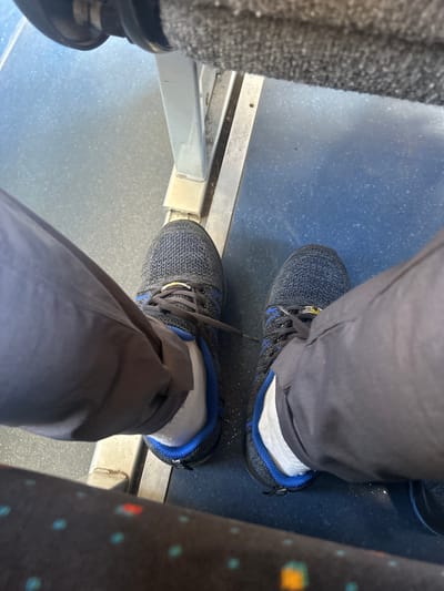 The image shows a close-up view of a person's legs and feet, likely from the perspective of someone sitting down. The person is wearing dark grey pants and dark-colored athletic shoes with white socks. The shoes have blue accents around the collar and laces. The person is sitting on a surface that appears to be a seat with a patterned fabric, possibly in a public transportation setting like a bus or train, given the presence of a metal railing and a textured floor. The floor is a dark blue speckled material. To the left of the frame, a grey, fuzzy textured material is visible, possibly the side of a seat or a bag. The lighting suggests it is daytime, and there are no indications of the weather. The scene is static, with no overt activity depicted, other than the implied act of sitting. No visible text is present in the image. The overall impression is one of everyday travel or waiting.