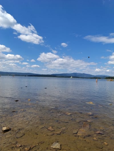 The image depicts a calm, wide lake under a bright blue sky with scattered white clouds. In the foreground, the shallow, clear water reveals a rocky bottom with numerous stones of varying sizes. The water's surface is gently rippled. In the middle ground, the lake extends to a distant, tree-covered shoreline with rolling hills and mountains beyond. Several sailboats are visible on the water, some with sails raised. On the right side of the image, a person is standing in the water. In the far distance, a yellow pedal boat with two occupants is present. A dark object, possibly a kite or a bird, is visible in the sky. The scene appears to be a leisure area, likely a lake or reservoir, with recreational activities taking place. The location is specified as Bližná, Czechia.
