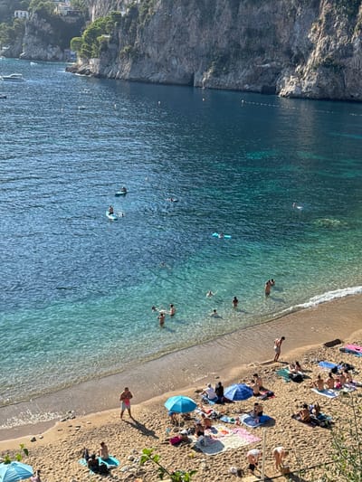 A high-angle forensic view captures Plage Mala, a beach and cove in Cap-d'Ail, France, under bright daylight. The scene encompasses a sandy beach, clear turquoise to deep blue water, and steep, vegetated rock cliffs forming a natural bay.

**Environment:**
The water, transitioning from shallow, transparent turquoise near the shore to a darker blue in deeper sections, reveals underwater rock formations. Gentle waves ripple along the sandy shoreline, which is interspersed with small pebbles. Towering, rugged cliffs with sparse green foliage define the bay's perimeter. Several structures, including residential buildings, are visible on the upper left cliff face.

**Subjects and Objects:**
Approximately 45-50 individuals are present.
*   **In the Water:** Roughly 17-20 people are engaged in water activities. Two individuals are on paddleboards; one stands, the other sits. One person floats on a light blue inflatable mat. The remaining individuals are swimming, wading, or floating close to the shore. Several small white boats and yachts are anchored further out in the bay. Yellow buoys and a segmented white rope barrier mark sections of the water.
*   **On the Beach:** Around 25-30 people occupy the light-colored sand. Most are spread out on beach towels or mats of various colors (pink, blue, patterned), primarily sunbathing or sitting. Three blue beach umbrellas provide shade. One individual stands near the waterline, facing the water, while another stands further back holding a pink item. Two individuals in the lower right are bent over, possibly adjusting items. Personal belongings like bags and bottles are visible near groups.

**Actions and Interactions:**
Individuals in the water are swimming, paddleboarding, and relaxing on an inflatable. On the beach, people are sunbathing, sitting, and standing. No explicit interactions between individuals are discernible due to the distance, but people are clustered in groups, suggesting social engagement. The overall scene depicts a busy recreational area.