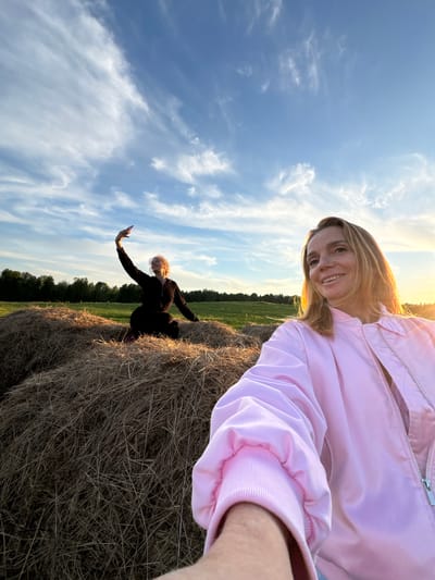 The image captures an outdoor scene in a rural setting, illuminated by the warm, golden light of late afternoon or early evening. The sky overhead is a bright blue with scattered white, wispy clouds, indicating clear weather.

In the foreground, a woman with long, light brown hair, wearing a vibrant pink jacket, is taking a selfie. She is smiling broadly towards the camera, her face and hair beautifully lit by the low sun. Her arm is visible on the right, extending to hold the camera.

Further back, positioned on a large, textured hay bale, another woman with lighter hair, dressed in dark clothing, is seen posing. She holds a smartphone in her outstretched hand, seemingly capturing a photo or video of the landscape or herself. The hay bales are prominent, made of dried straw. Beyond them, a green field extends towards a distant treeline. The overall mood is cheerful and relaxed, depicting an enjoyable outdoor moment in the region of Стеклянный, Russia. No visible text is present in the image.