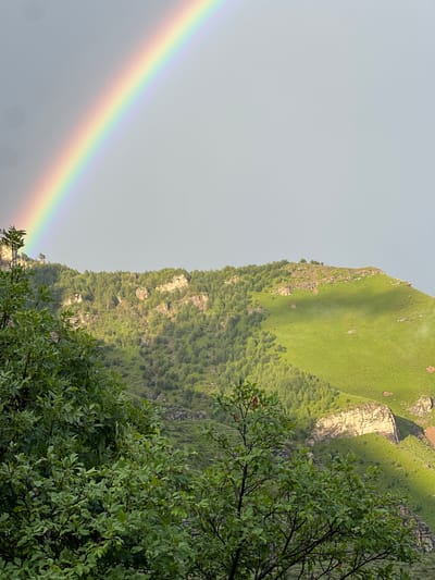 This is a photograph of a mountainous landscape with a rainbow in the sky. In the foreground, there are green leafy trees. Behind the trees, a lush green mountain rises, covered with trees on its slopes and rocky outcrops. A vibrant rainbow arches across the grey, overcast sky, its colors clearly visible. The scene appears to be natural and uninhabited, with no people or man-made structures visible. The lighting suggests it may have recently rained or is about to. The location is stated to be in the city of Assab, Russia, a detail that cannot be verified from the image itself.