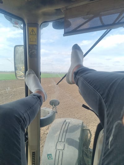 The image presents an interior view from a tractor cab, looking outward over agricultural fields near Svoboda, Bulgaria. Two human legs, clad in dark grey jeans and light grey ankle socks, are elevated and rest against the interior frame of the cab, crossing the central vertical pillar and extending towards the right.
The interior of the cab shows a beige-colored frame and ceiling. A warning sticker on the central pillar displays a yellow triangle with an exclamation mark symbol above an open book icon. A rectangular mesh sunshade is partially visible and retracted at the top of the windshield. A side-view mirror is visible on the left, reflecting a yellow agricultural implement in the distance. Part of a dark grey tractor fender with a yellow John Deere logo is visible through the window on the lower right. An exterior work light is mounted below the left leg.
The exterior environment, viewed through the cab windows, features a vast expanse of brown, tilled soil in the foreground. Further in the distance, a strip of green vegetation is visible, leading to a linear formation of trees along the horizon. The sky above is partly cloudy with significant patches of blue. The overall scene suggests a pause in agricultural activity within a rural landscape.