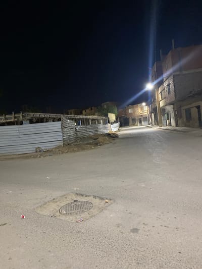 A street scene in Sidi Chahmi, Algeria at night. The sky is black, the scene dimly lit by a street light. Buildings line the left side of the street. A metal corrugated fence is adjacent to the street, alongside a building construction site. A manhole cover is visible on the pavement.