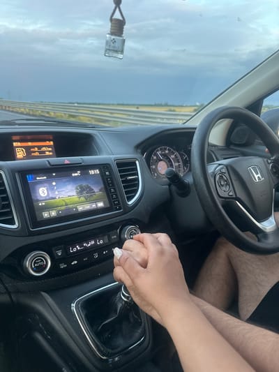 The image captures the interior of a car, likely a Honda given the steering wheel logo, from a slightly overhead perspective. Two individuals are present: one person's hairy legs are visible near the steering wheel, and another person's hand, with white nail polish, is holding the first person's hand on the gear shifter.
The scene appears to be set during the evening, indicated by the time "18:15" (6:15 PM) displayed on the car's infotainment system. The sky visible through the windshield is overcast with a light blue-grey hue, suggesting either a cloudy day or approaching dusk. A road with multiple lanes or structures runs parallel to the horizon in the background. A small air freshener labeled "BLUIT J" hangs from the rearview mirror.
The car's dashboard features a digital infotainment screen showing music playback, with "iPhone" and "Bluetooth" visible, and track controls. The current music is track "16" out of "4" (likely a display error or segment of a long track). To the left of the screen, another display shows "AVG 19.1 mpg" and "AVG 38 mi". The climate control panel has buttons for "LO" temperature, "SYNC", "A/C", "FRONT", and "REAR", with a "PUSH AUTO" button for automatic climate control. The instrument cluster shows speed markers up to "120" (likely mph or km/h). The overall setting is inside a moving or stationary vehicle, possibly in Makó, Hungary, depicting a moment of connection between two individuals.
