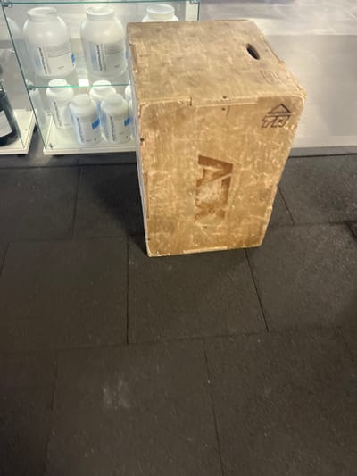 A worn, light-brown wooden plyometric box, featuring a prominent "ATX" logo and a geometric symbol in the upper right corner, stands on a dark gray interlocking rubber tiled floor. A circular handle cutout is visible on the box's top surface. To the left, a clear glass display case exhibits multiple white plastic supplement containers, some bearing "nutrimuscle" branding with blue labels. A dark-colored bottle is partially visible on a lower shelf within the display case. The background behind the display unit is a light gray wall. The scene is located within a fitness facility or supplement retail store in Wolfisheim, France. No individuals are present.