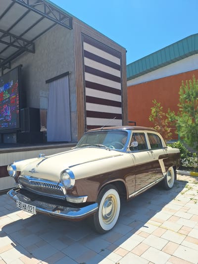 A vintage, two-tone beige and brown sedan is parked on a paved area in front of a building. The car has chrome accents, whitewall tires, and a hood ornament. Behind the car, a building features a large screen displaying colorful graphics, speakers, and a white curtain. The building has a patterned facade with horizontal brown and white stripes. To the right of the building, there is a section of orange wall, a green metal roof, and some evergreen trees. The sky is clear and blue. The license plate on the car reads "90 HA 021". The scene appears to be outdoors, in what could be a public space or event venue in Khudat, Azerbaijan.