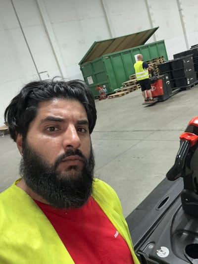 A man with dark hair and a beard, wearing a red shirt and a bright yellow safety vest, takes a selfie in what appears to be a warehouse environment. In the background, another man in a similar yellow vest is operating a pallet jack near a large green dumpster. The floor is concrete, and the walls are white. The scene suggests a commercial or industrial setting.