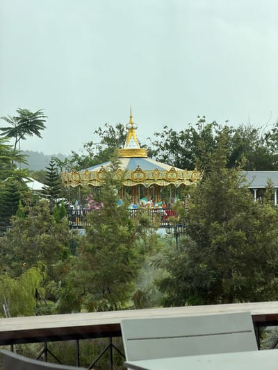 The image, taken in the vicinity of Ban Samnak Tabaek, Thailand, shows a carousel situated amidst lush greenery. The carousel features a golden roof with intricate blue and gold detailing, and is adorned with decorative gold elements. Various colorful horses and figures are visible on the carousel's platform, suggesting it is in operation. Surrounding the carousel are numerous trees and foliage, including tall, conical evergreen trees and some deciduous trees with lighter green leaves. In the background, a light-colored building with a gray roof and white trim can be seen, partially obscured by trees. The sky is overcast with a pale, uniform gray color. In the foreground, a wooden table and part of a white chair are visible, suggesting a seating area with a view of the carousel and garden. The overall impression is of a serene, park-like setting.
