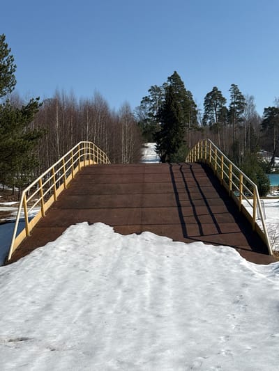 This image captures a pedestrian bridge in Oleshkovo, Russia, under clear skies. The bridge features a dark brown, textured metal surface that arches upwards slightly towards the center. Bright yellow or cream-colored metal railings, each with multiple horizontal bars, line both sides of the bridge, curving to match its profile. Strong sunlight illuminates the scene, casting distinct shadows of the right-side railing onto the bridge's surface.
The immediate ground surrounding the bridge is covered in patchy white snow, indicating a winter or early spring setting. Footprints are visible in the snow in the foreground, approaching the bridge from the left. Sparse brown earth and dead vegetation are visible where the snow has melted.
Dense tree lines flank the bridge and extend into the background. Evergreen conifer trees, primarily pines, are prominent on both sides of the foreground. Behind them, a mix of deciduous trees with bare branches and additional conifers form a deeper forest. A light-colored, unpaved path or road is discernible winding uphill through the trees in the distant background, aligning with the bridge's trajectory. The sky above is clear and bright blue, devoid of clouds. No individuals or active subjects are present in the frame.