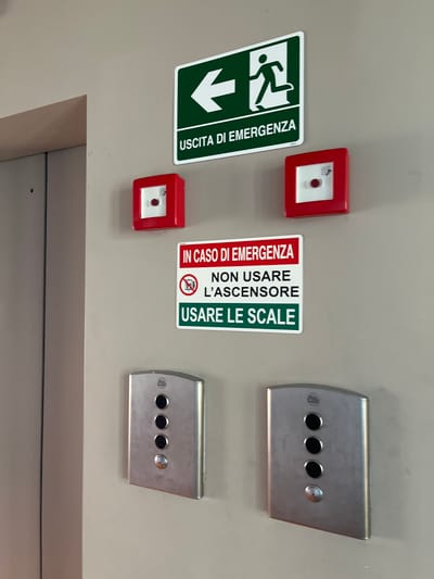 This media file captures an indoor scene, likely a hallway or area adjacent to an elevator in a building in Dorno, Italy. Mounted on a plain, light-colored wall are various safety and emergency components.
To the left, a green emergency exit sign displays a pictogram of a running person and a downward-pointing arrow, accompanied by the Italian text "USCITA DI EMERGENZA" (Emergency Exit).
In the center, two red square-shaped emergency break glass units are visible, positioned vertically. Between these red units and to their right, a white rectangular sign with red and green sections provides emergency instructions. The red section at the top reads "IN CASO DI EMERGENZA" (In Case of Emergency). Below this, the white section features text "NON USARE L'ASCENSORE" (Do Not Use the Elevator) alongside a crossed-out elevator pictogram. The bottom green section instructs "USARE LE SCALE" (Use the Stairs).
To the right, two identical metallic control panels are mounted vertically. Each panel has a curved, brushed metal finish and features three black circular buttons and one smaller, circular silver button or indicator light. These panels are typical elevator call buttons.
The image is taken from a slight diagonal angle, making the mounted items appear tilted on the wall. No specific activity is occurring, as the image shows fixed installations. The visible Italian text and the nature of the signs strongly suggest a public or commercial building setting, designed to guide occupants during an emergency.