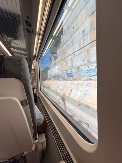 The image shows the interior of a moving train, looking out the window at a concrete retaining wall with graffiti. The seats are visible in the foreground, along with part of a passenger's legs. The retaining wall is weathered and shows signs of damage, with vegetation growing in crevices. The graffiti is spray-painted in various colors and styles. The train appears to be traveling through a rural or industrial area, as evidenced by the retaining wall and the general lack of other structures visible outside. The scene suggests a journey through a landscape that has been altered by human activity. There is no indication that this location is in the city of Cabanes, Spain.