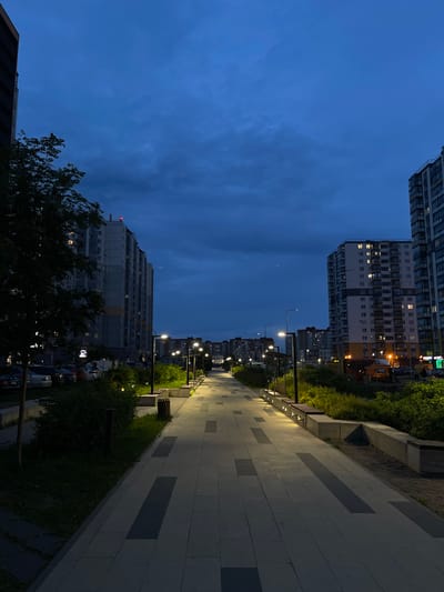 The image captures a view of a modern residential area in Novogorelovo, Russia, during twilight. A paved pedestrian walkway with embedded lights runs through the center of the frame, bordered by green spaces and benches. Tall apartment buildings with illuminated windows line both sides of the walkway, indicating residential activity. The sky is a deep, cloudy blue, characteristic of dusk. Cars are parked along the side of the road to the left, and some streetlights illuminate the path. A large tree is present on the left side of the walkway, casting a shadow.