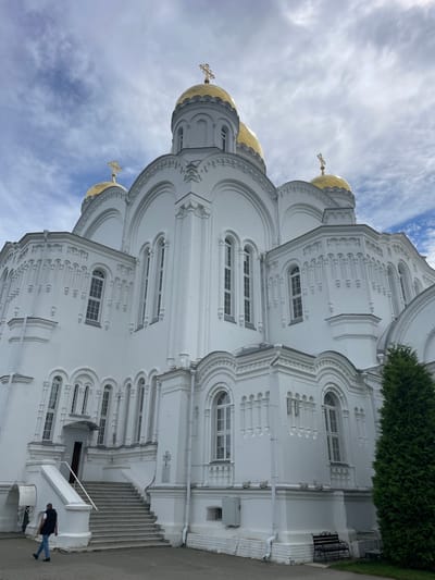 The image shows a low-angle view of a large, white, Orthodox church with golden domes, located in Diveyevo, Russia. The church has intricate architectural details, including numerous arched windows and decorative carvings. The sky is partly cloudy, with patches of blue visible. In the foreground, a man in a dark vest and blue jeans is walking down a wide set of stairs leading to an entrance of the church. To the right of the stairs, a tall, green coniferous tree stands prominently. A black bench and a silver trolley are visible near the base of the church. The ground is paved with grey stone. The scene appears to be during daylight.
