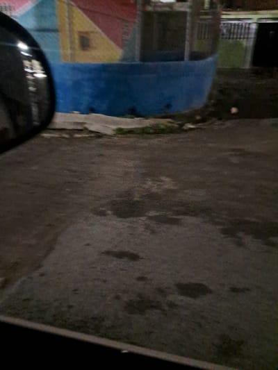 The image depicts a side-view mirror reflecting a colorful mural painted on a blue, curved wall. The mural features geometric shapes in pink, yellow, and red. To the right of the blue wall, there is a glimpse of buildings with metal fencing and green vegetation. The foreground shows a dark, wet asphalt surface with several puddles reflecting the ambient light. The scene appears to be an outdoor urban environment in Cumana, Venezuela, likely at dusk or night, given the lighting conditions and the reflections. There are no people visible in the image.
