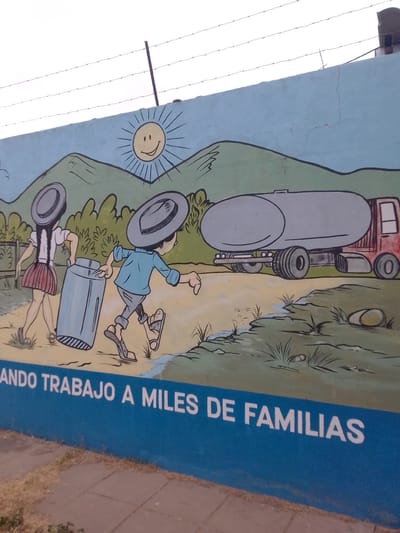 A painted mural on a wall depicts a rural scene with cartoon-like figures. A smiling sun is painted in the sky above green hills. A boy and a girl wearing hats are carrying a container toward a red truck with a grey tank. The environment is grassy with a small stream. Barbed wire is visible along the top of the wall. The phrase "ANDO TRABAJO A MILES DE FAMILIAS" is painted in white letters on a blue stripe at the bottom of the wall. The location is Municipio Tarija, Bolivia. A sidewalk is visible in the foreground.