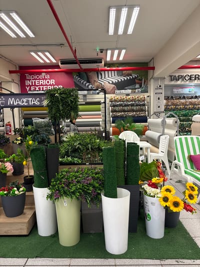 This is a well-lit, eye-level medium shot of a retail store display, likely in La Victoria, Peru, featuring various artificial plants, planters, and fabrics. The store appears to be a fabric and home decor store, with rolls of fabric stacked vertically in the background and signage indicating "Tapicería INTERIOR Y EXTERIOR" (Upholstery INTERIOR AND EXTERIOR) and "MACETAS" (Pots).
In the foreground, several large, cylindrical planters are arranged on a green artificial turf floor. Two are white, one is light green, and one is dark gray. These planters contain artificial plants of varying types and colors. To the left, a dark gray planter holds a small artificial tree with yellow lemons and some pink flowers in a black pot. Next to it, a white planter is filled with green bushes and purple flowers. A light green planter overflows with dense green foliage and smaller purple flowers. To the right, a white planter holds an arrangement of artificial sunflowers and red flowers.
Two tall, dark green cylindrical objects, possibly rolls of artificial grass or matting, stand in front of the planters. Behind these, a variety of artificial plants are displayed, including lush green bushes, ferns, and flowering plants.
The midground reveals a display of furniture, including white plastic chairs and a small table. Further back, on shelves, are numerous rolls of patterned and solid-colored fabrics, suggesting the "Tapicería" aspect of the store. A woman wearing a gray jacket is partially visible in the background, looking towards the right.
The environment is indoors, with bright fluorescent lighting on the ceiling. The store appears to be a commercial establishment with a focus on home furnishings and outdoor decor. The overall impression is one of abundance and variety, showcasing decorative items for both interior and exterior spaces.
