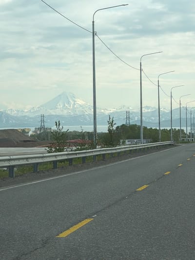An eye-level, vertical medium shot captures a road leading towards a snow-capped mountain range under a cloudy sky. The road, surfaced with asphalt, features broken yellow lines as lane markers. On the left, a silver metal guardrail runs along the roadside, with a grassy embankment behind it. In the middle ground, several electrical transmission towers are visible, along with a body of water and lush green trees. A large, conical, snow-covered mountain dominates the background, with smaller, snow-capped peaks extending to its sides. The scene is likely located in or near the city of Svetly, Russia, given the prominent mountainous landscape.