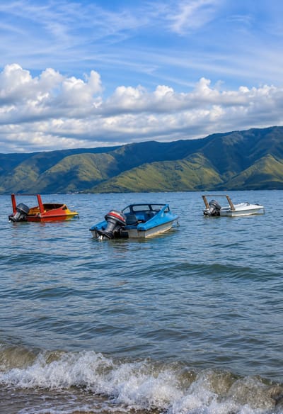 Three small motorboats are moored on a body of water, likely Lake Toba, near the shore of Situngkir, Indonesia. The boats are positioned at varying distances from the viewer, with two closer to the foreground and one further back. One boat is bright red and yellow, another is blue and white, and the third is white with wooden trim. All three have outboard motors. In the background, lush green mountains rise up, meeting a bright blue sky filled with scattered white clouds. Gentle waves lap against a sandy shore in the foreground. The scene is serene and captures a typical coastal landscape.
