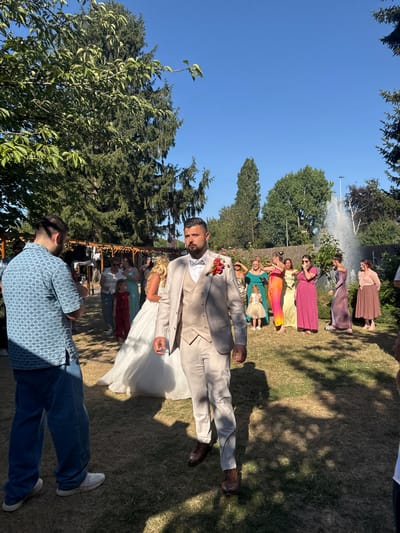 The image depicts an outdoor wedding ceremony in Hem, France. The scene is set on a grassy area surrounded by trees and features a fountain in the background. In the foreground, a groom in a light tan suit with a bow tie and a boutonniere is looking to his right. To his left, a man in a blue patterned shirt and jeans is holding a video camera. Several women in colorful dresses and a young girl in a red outfit are gathered in the midground, observing the ceremony. A bride in a white wedding dress is also visible, facing the groom. The atmosphere suggests a celebration with guests and a professional videographer capturing the event. The lighting indicates it is daytime, with shadows cast by the trees on the grass.