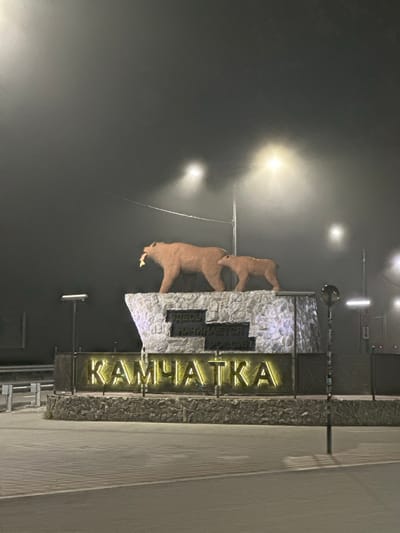 The image shows a monument dedicated to Kamchatka, located in the city of Pionersky, Russia. The monument features two bear sculptures, one adult and one cub, atop a stone structure. Inscribed on the stone are the words "ЗДЕСЬ НАЧИНАЕТСЯ РОССИЯ" (Here begins Russia). Below the stone base, illuminated in neon, are the letters "КАМЧАТКА" (Kamchatka). The scene is set at night, with streetlights and fog creating a hazy atmosphere. The ground in front of the monument appears to be paved.