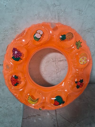 A close-up, overhead shot shows an inflatable orange swim ring with various fruit images printed on it. The ring is laid flat on a textured, grey surface. The fruits depicted include grapes, strawberries, bananas, watermelon, cherries, oranges, and a pineapple. The ring has a transparent quality, allowing the texture of the surface underneath to be partially visible. There are some small air valves visible on the surface of the ring. The background is a grey, textured wall. The location of this image in Қанабек, Kazakhstan, is not discernible from the visual information provided.