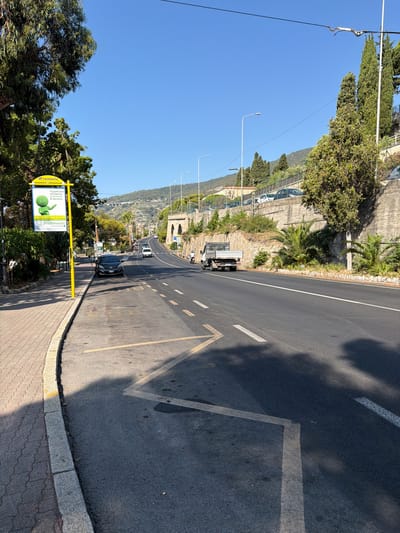 The image depicts a street scene in Ospedaletti, Italy, on a sunny day. The foreground shows a sidewalk with a textured paving pattern bordering a road with various markings, including yellow dashed lines and white directional arrows. A dark-colored car is parked along the curb. A yellow bus stop sign with an advertisement is visible on the left side of the road. In the mid-ground, the road curves upwards, flanked by a stone retaining wall covered with vegetation. Several parked cars are visible on a higher level behind a fence. A white flatbed truck is driving away from the camera on the road. The background features rolling hills with buildings and trees, and a clear blue sky. The overall environment is urban with natural elements.