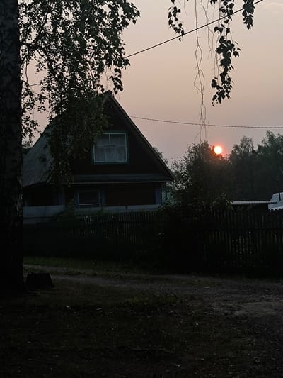 This media file captures a rural scene in Maiskii, Russia, during sunset. The primary subjects are a traditional wooden house with a steep roof and a weathered wooden fence. The house appears to be a dacha, a common type of summer house or cottage in Russia. Hanging branches with leaves, likely from a birch tree given the bark visible on the left, frame the upper portion of the image. In the background, a dense line of trees forms a dark silhouette against the hazy, orange-tinged sky.
The sun is a vibrant, glowing orb low on the horizon, partially obscured by the trees, indicating the time of day is sunset. The sky suggests a warm, possibly humid atmosphere, with the haze potentially caused by dust, smoke, or humidity. The overall mood is serene and peaceful, evoking a sense of quiet twilight in the countryside. The foreground shows a dark, unpaved path and grassy areas, with a tree stump visible near the large tree trunk on the left. The scene is devoid of any people or overt activity, emphasizing the stillness of the moment. No visible text is present in the image.